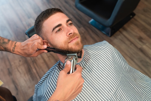 Photo gratuite jeune coiffeur rasant la barbe du client masculin avec un peigne et une tondeuse à cheveux dans un salon de coiffure