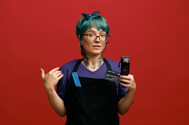 Jeune Coiffeuse Concentrée Portant Des Lunettes Uniformes Et Un Bandeau Montrant Une Carte De Crédit Et Une Tondeuse à Cheveux Regardant La Caméra Pointant Vers Elle-même Avec La Main Avec Un Peigne Dans Sa Poche