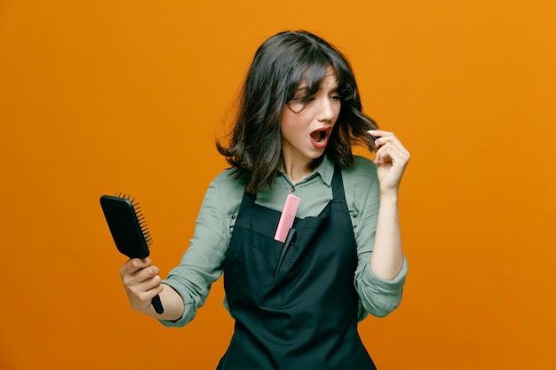 Jeune coiffeuse femme portant un tablier tenant une brosse à cheveux se peignant les cheveux l'air confus et inquiet debout sur fond orange