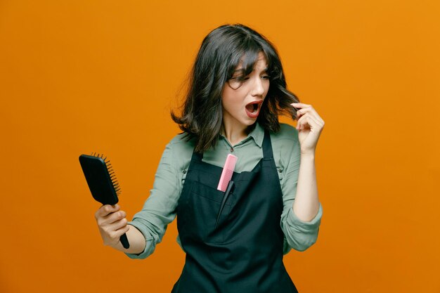 Jeune coiffeuse femme portant un tablier tenant une brosse à cheveux se peignant les cheveux l'air confus et inquiet debout sur fond orange