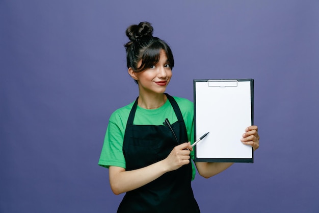 Jeune coiffeuse femme portant un tablier tenant un presse-papiers et un stylo regardant la caméra avec une expression confiante souriant debout sur fond bleu