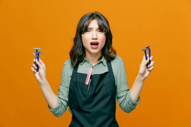 Jeune coiffeuse femme portant un tablier tenant des rasoirs regardant la caméra confuse et déçue debout sur fond orange