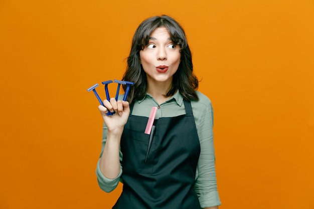 Jeune coiffeuse femme portant un tablier tenant des rasoirs regardant de côté avec une expression confuse debout sur fond orange