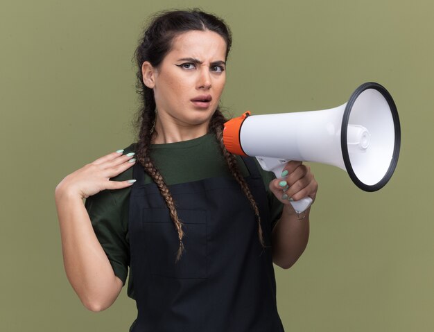Photo gratuite une jeune coiffeuse mécontente en uniforme parle sur un haut-parleur mettant la main sur l'épaule isolée sur un mur vert olive