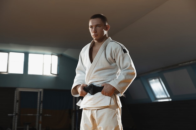 Photo gratuite jeune combattant de judo en kimono posant confortablement dans la salle de gym forte et saine