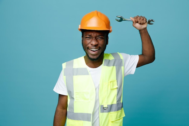 Jeune constructeur afro-américain en colère en uniforme tenant une clé à fourche isolée sur fond bleu