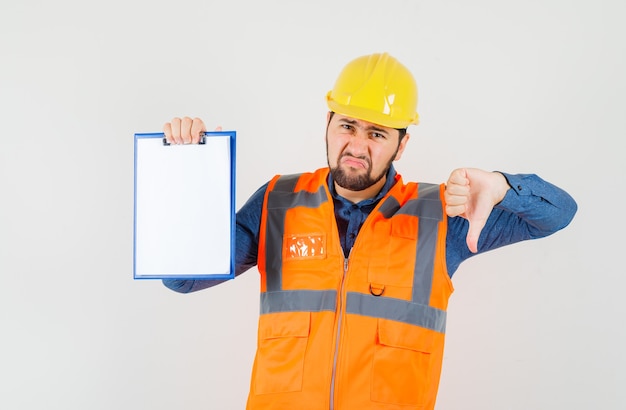 Jeune constructeur en chemise, gilet, casque montrant le pouce vers le bas, tenant le presse-papiers et regardant mécontent, vue de face.