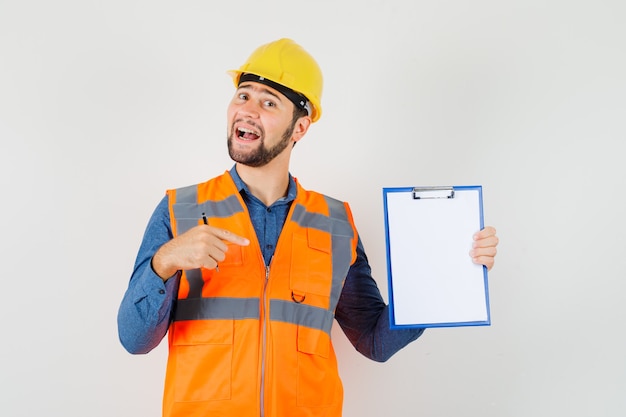 Jeune Constructeur En Chemise, Gilet, Casque Pointant Sur Le Presse-papiers Et à La Joyeuse Vue De Face.