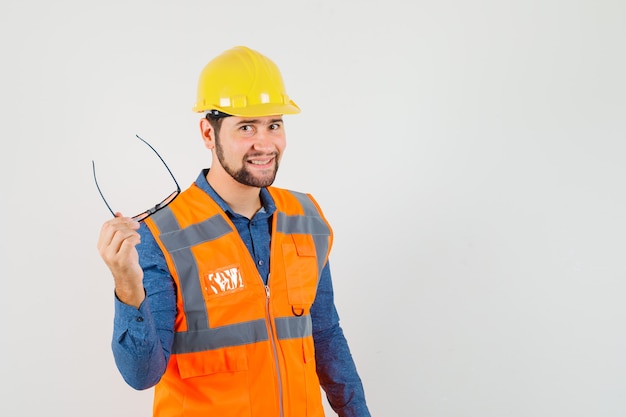 Jeune constructeur en chemise, gilet, casque tenant des lunettes et regardant gai, vue de face.