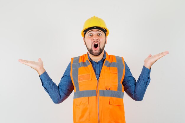 Jeune constructeur faisant le geste des échelles en chemise, gilet, casque et à la surprise, vue de face.