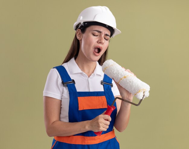 Jeune constructeur femme aux yeux fermés en uniforme tenant la brosse à rouleau et chant isolé sur mur vert olive