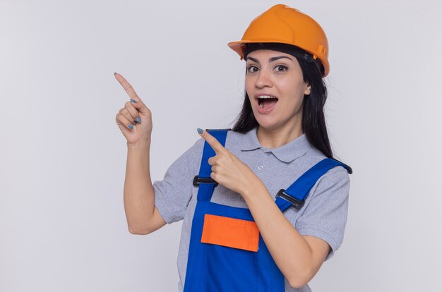 Jeune constructeur femme en uniforme de construction et casque de sécurité à l'avant souriant heureux et positif pointant avec l'index sur le côté debout sur un mur blanc