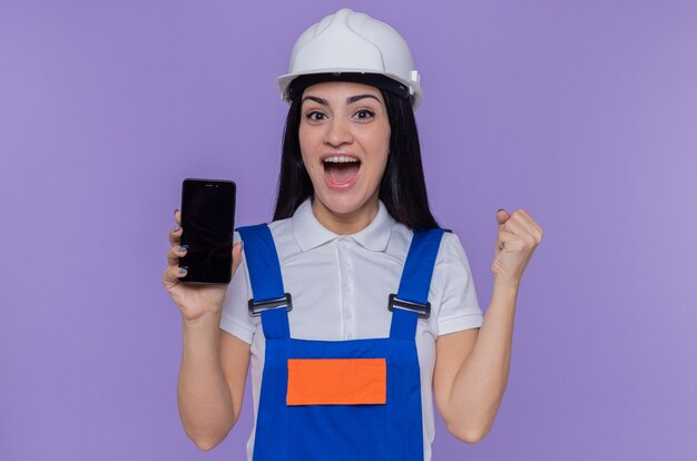 Jeune constructeur femme en uniforme de construction et casque de sécurité montrant smartphone à l'avant poing serré heureux et excité debout sur le mur violet