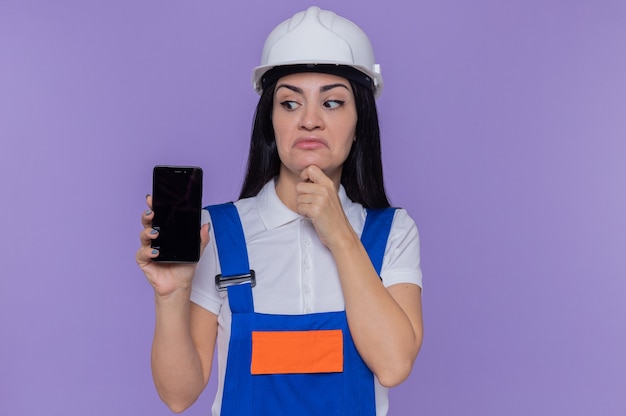 Jeune constructeur femme en uniforme de construction et casque de sécurité montrant smartphone en le regardant avec la main sur le menton avec une expression pensive pensant debout sur le mur violet