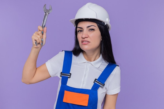 Jeune constructeur femme en uniforme de construction et casque de sécurité tenant une clé à l'avant avec une expression sceptique debout sur un mur violet