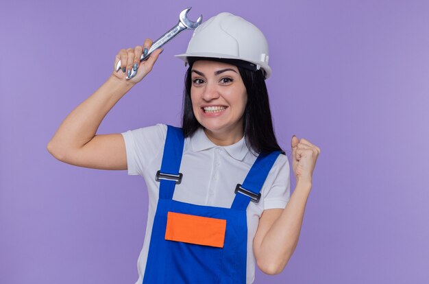 Jeune constructeur femme en uniforme de construction et casque de sécurité tenant la clé serrant le poing heureux et excité debout sur le mur violet