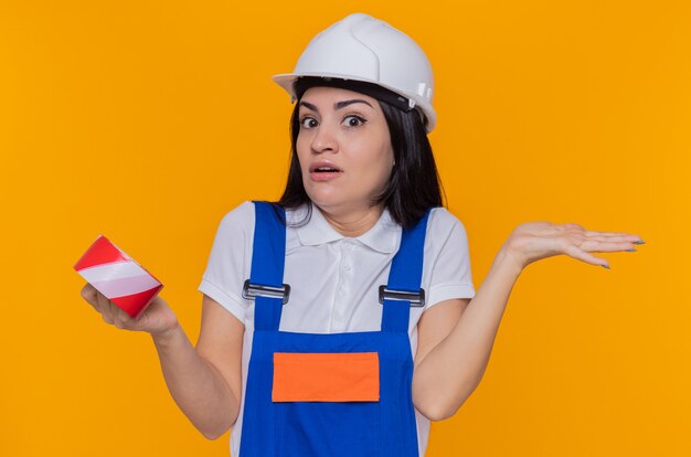 Jeune constructeur femme en uniforme de construction et casque de sécurité tenant du ruban adhésif à l'avant en haussant les épaules confus ayant des doutes debout sur le mur orange