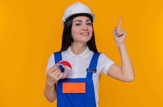 Jeune constructeur femme en uniforme de construction et casque de sécurité tenant du ruban adhésif souriant confiant montrant l'index debout sur le mur orange