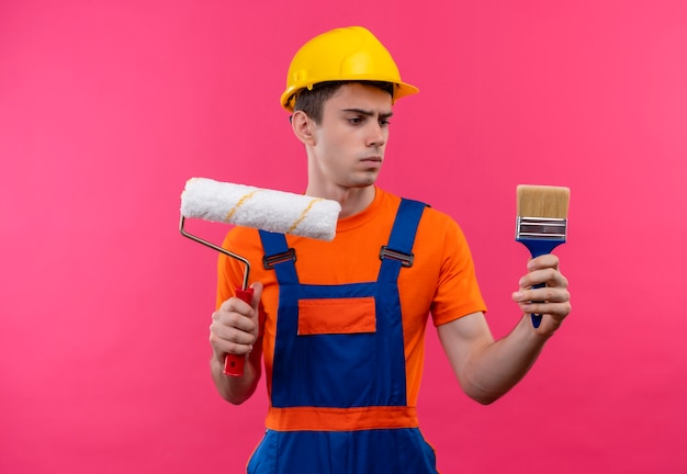 Jeune Constructeur Homme Portant L'uniforme De Construction Et Un Casque De Sécurité Détient Un Pinceau à Rouleau Et Un Pinceau Mural