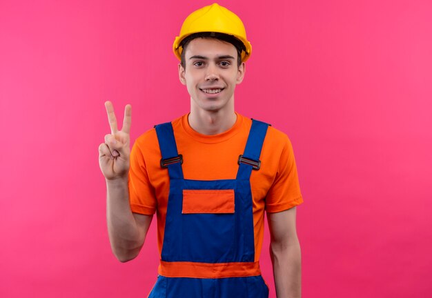 Jeune constructeur homme portant des uniformes de construction et un casque de sécurité sourit et montre un geste de paix