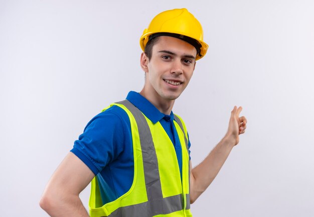 Jeune constructeur homme portant des uniformes de construction et un casque de sécurité sourit et montre en plus