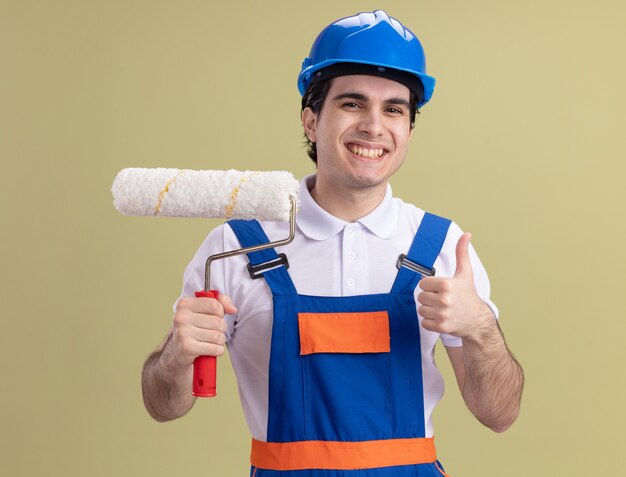 Jeune constructeur homme en uniforme de construction et casque de sécurité tenant le rouleau à peinture à l'avant heureux et positif montrant les pouces vers le haut debout sur le mur vert