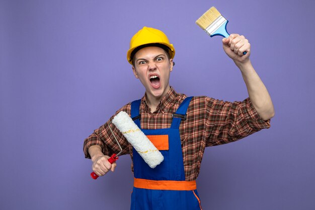 Jeune constructeur masculin portant l'uniforme tenant une brosse à rouleau avec un pinceau isolé sur un mur violet