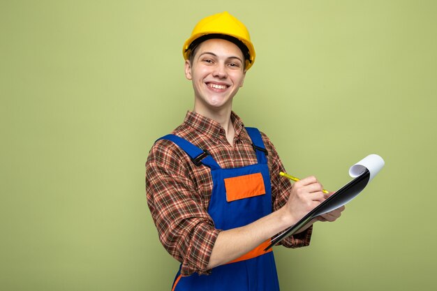 Le jeune constructeur masculin de sourire écrit sur le presse-papiers portant l'uniforme