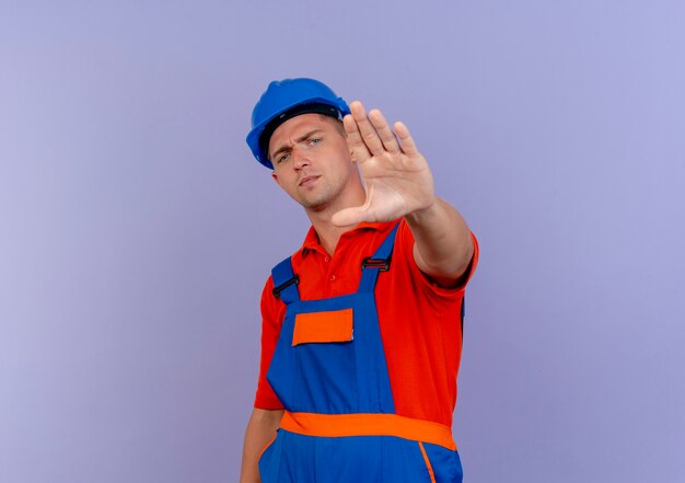 Jeune constructeur masculin strict portant l'uniforme et un casque de sécurité montrant le geste d'arrêt sur violet