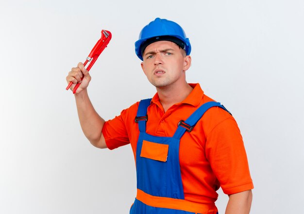 Jeune constructeur masculin strict portant l'uniforme et un casque de sécurité soulevant une clé à gaz sur blanc
