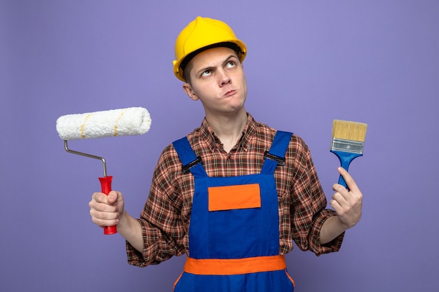 Jeune constructeur masculin en uniforme tenant un pinceau avec une brosse à rouleau isolée sur un mur violet