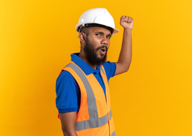 Photo gratuite jeune constructeur mécontent en uniforme avec un casque de sécurité levant le poing isolé sur un mur orange avec espace de copie