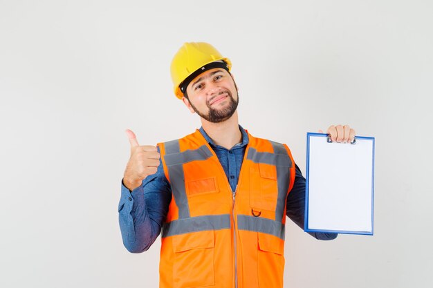 Jeune constructeur montrant le pouce vers le haut, tenant le presse-papiers en chemise, gilet, casque et regardant joyeux, vue de face.