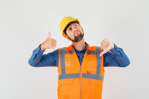 Jeune constructeur montrant les pouces de haut en bas en chemise, gilet, casque et à l'hésitation. vue de face.