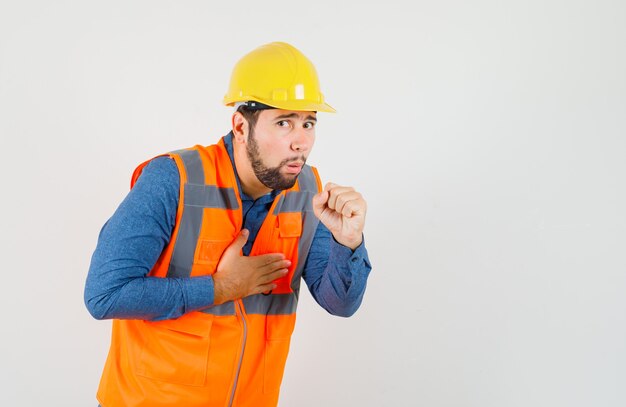 Jeune constructeur souffrant de toux en chemise, gilet, casque et à la recherche de mal. vue de face.