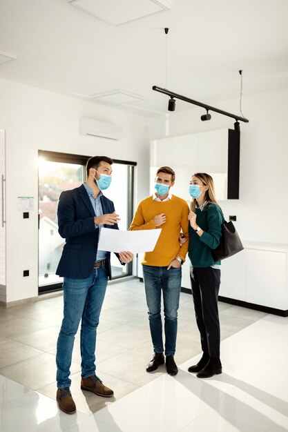 Jeune couple achetant une nouvelle maison et examinant les plans avec un agent immobilier tout en portant des masques faciaux