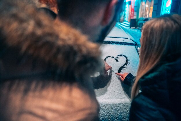 Jeune couple adulte dessine un coeur sur une voiture couverte de neige