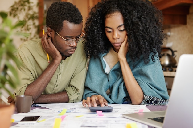 Un jeune couple africain stressé ne supporte pas la tension de la crise financière, l'air malheureux et frustré, assis à la table de la cuisine avec une calculatrice, essayant d'économiser de l'argent en réduisant les dépenses domestiques