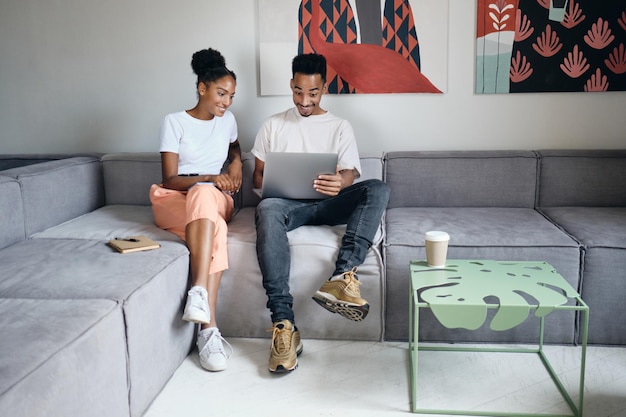 Jeune couple afro-américain décontracté attrayant travaillant joyeusement sur un ordinateur portable ensemble sur un canapé dans une maison moderne