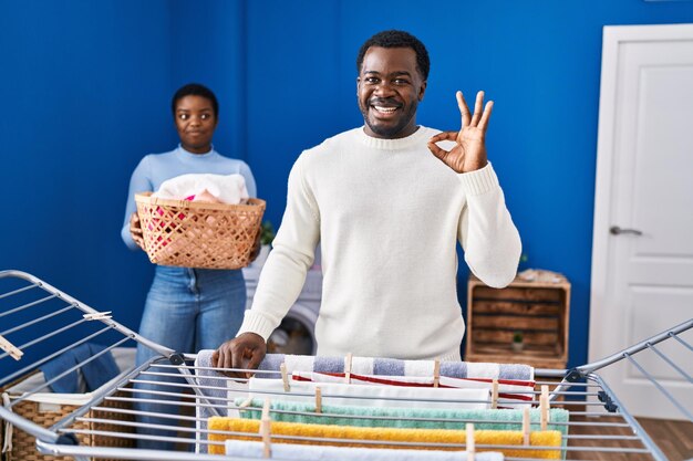 Jeune couple afro-américain vêtements suspendus à la corde à linge faisant signe ok avec les doigts souriant amical gesticulant excellent symbole