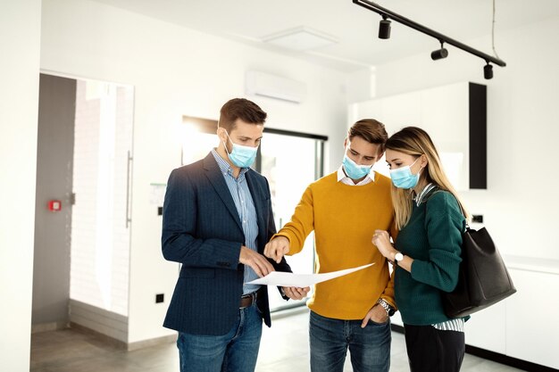 Jeune couple et agent immobilier portant des masques faciaux lors de l'analyse des plans de logement