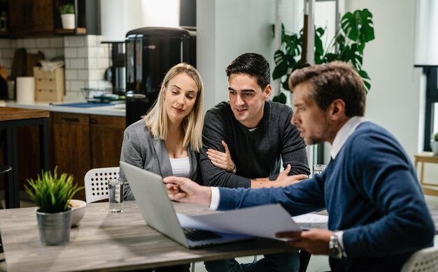 Jeune couple et agent immobilier utilisant un ordinateur portable tout en examinant le plan de logement lors d'une réunion