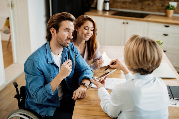 Jeune couple et agent immobilier utilisant un pavé tactile lors d'une réunion à la maison L'accent est mis sur l'homme en fauteuil roulant