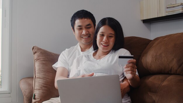 Jeune couple asiatique enceinte achats en ligne à la maison. Maman et papa se sentent heureux d'utiliser un ordinateur portable et une carte de crédit pour acheter un produit pour bébé en position allongée sur le canapé du salon à la maison.