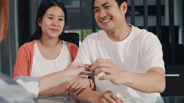 Un jeune couple asiatique enceinte signe des documents contractuels à la maison, une famille japonaise consulte un conseiller financier en immobilier, l’achat d’une nouvelle maison et la négociation avec un courtier remettant les clefs dans le salon.