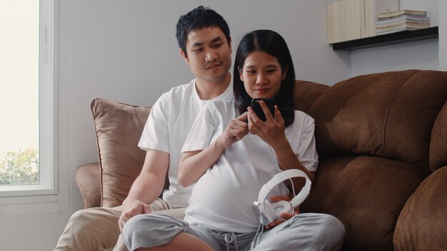 Jeune couple asiatique en utilisant un téléphone et un casque joue de la musique pour bébé dans le ventre. Maman et papa se sentent heureux, souriant et serein tout en prenant soin d’enfant couché sur un canapé dans le salon à la maison