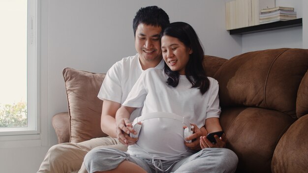 Jeune couple asiatique en utilisant un téléphone et un casque joue de la musique pour bébé dans le ventre. Maman et papa se sentent heureux, souriant et serein tout en prenant soin d’enfant couché sur un canapé dans le salon à la maison
