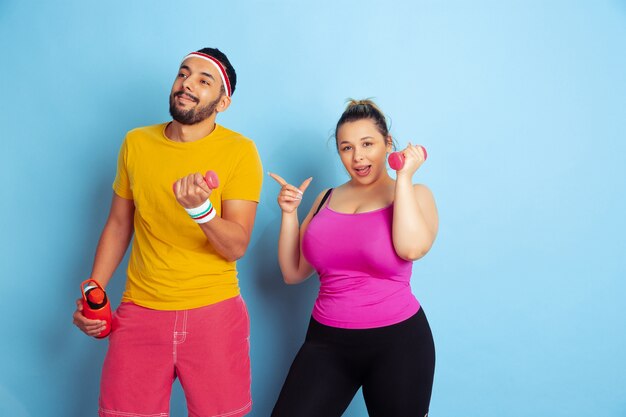 Jeune couple assez caucasien en formation de vêtements lumineux sur fond bleu Concept de sport, émotions humaines, expression, mode de vie sain, relation, famille. Entraînement avec des poids, amusez-vous.