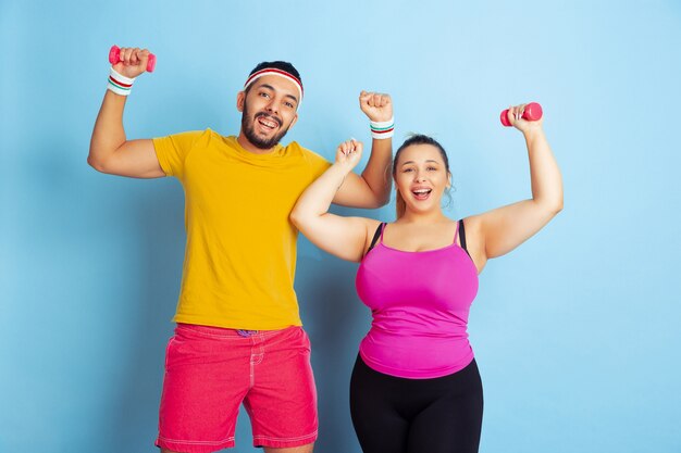 Jeune couple assez caucasien en formation de vêtements lumineux sur fond bleu Concept de sport, émotions humaines, expression, mode de vie sain, relation, famille. Entraînement avec des poids, amusez-vous.