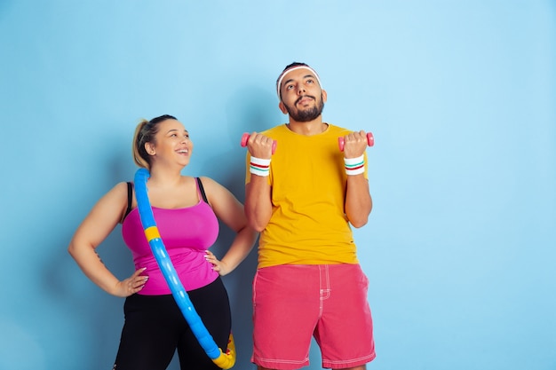 Jeune couple assez caucasien en formation de vêtements lumineux sur fond bleu Concept de sport, émotions humaines, expression, mode de vie sain, relation, famille. Pratiquer avec cerceau et poids.